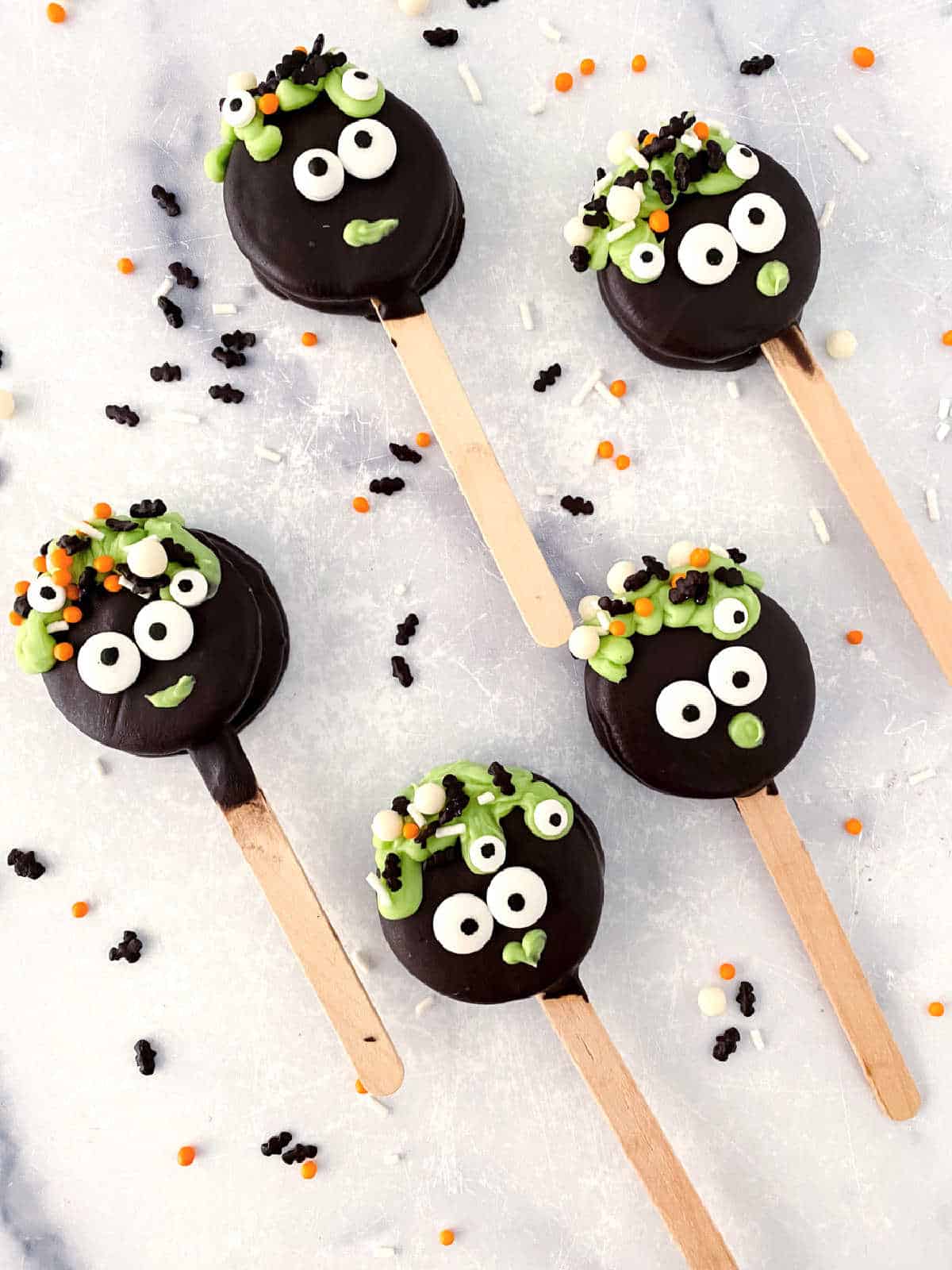 Five Halloween-themed chocolate cake pops decorated with googly eyes and green, orange, and black icing faces on wooden sticks, reminiscent of bubbling cauldrons. They rest on a white surface with scattered sprinkles.