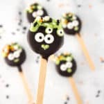 Four Halloween-themed chocolate popsicles, decorated to look like monsters with googly eyes and green icing on wooden sticks, resembling delightful cauldron pops.