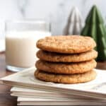 Four stacked cookies on a book with a glass of milk and decorative trees in the background.
