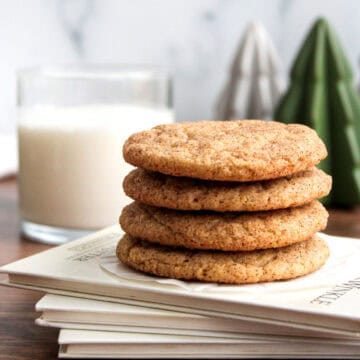Four stacked cookies on a book with a glass of milk and decorative trees in the background.