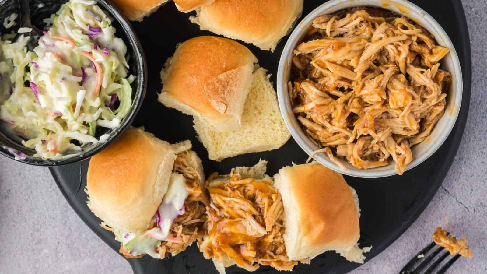A plate with pulled chicken sliders on buns, a bowl of coleslaw, and a bowl of pulled chicken.