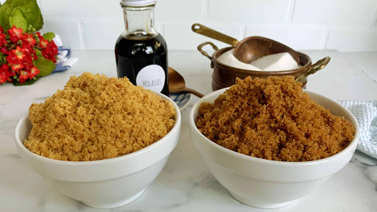 Two bowls of brown sugar are placed on a countertop with a bottle of molasses, a bowl of white sugar, a copper saucepan, and a red flower in the background.