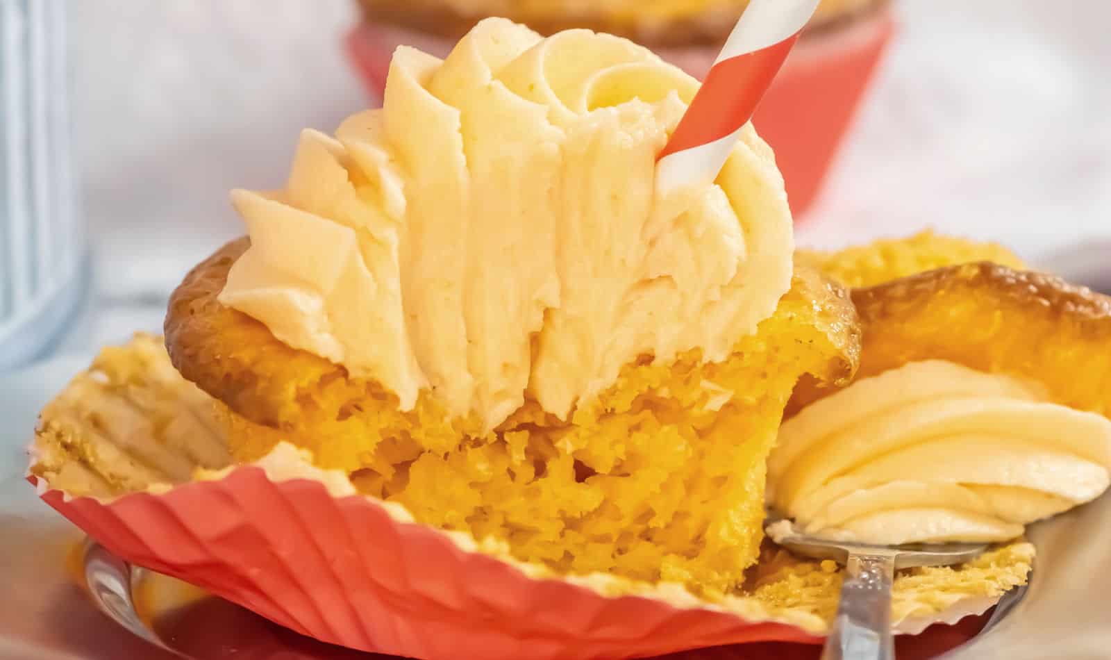 A cupcake with yellow cake and a generous swirl of white frosting, partially unwrapped from its red liner, with a red and white straw inserted in the frosting.