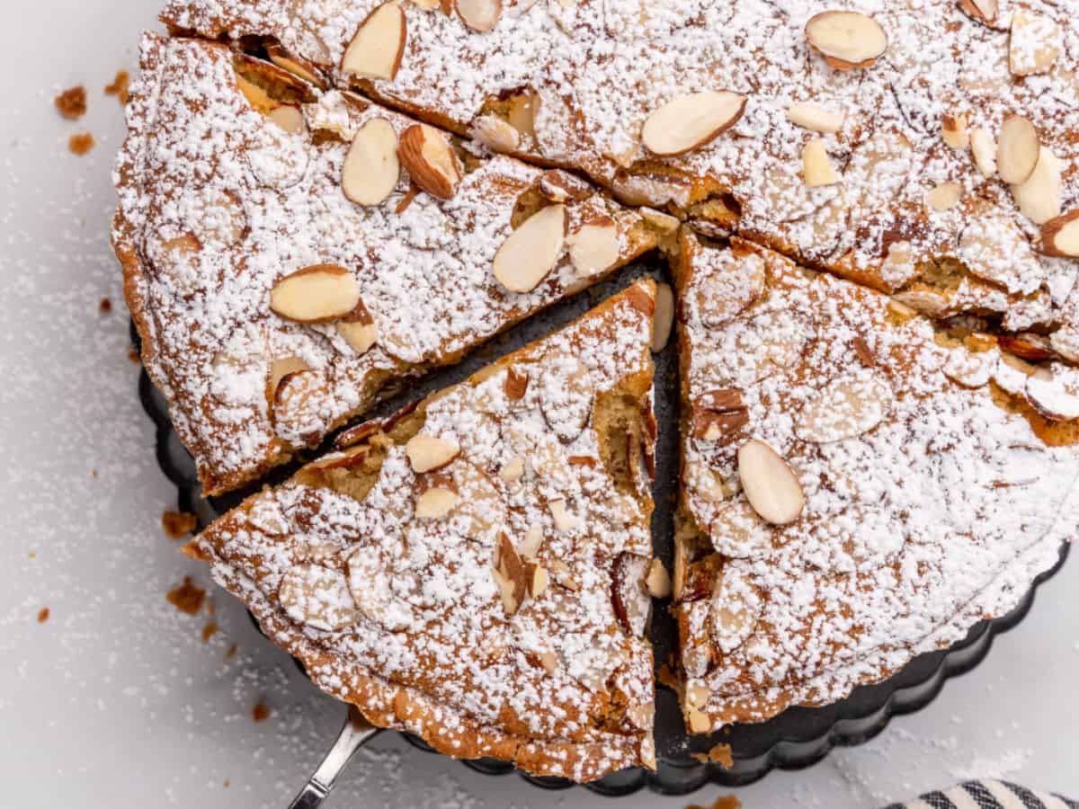 A round almond cake dusted with powdered sugar is cut into slices. It sits on a serving plate with a portion being lifted.