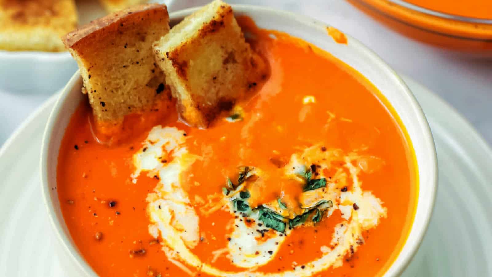 A bowl of tomato soup garnished with a swirl of cream, herbs, and two pieces of toasted bread.