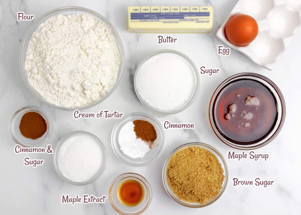 Top-down view of various cookie ingredients labeled on a white surface, including flour, sugar, butter, egg, cinnamon, maple syrup, brown sugar, cream of tartar, and maple extract.