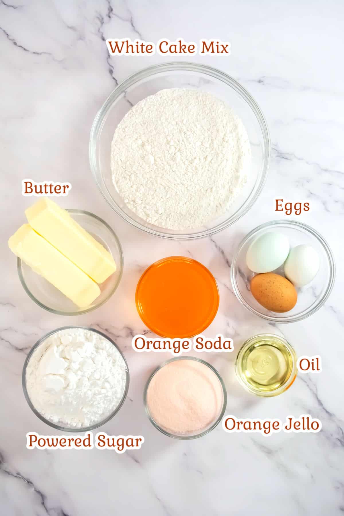 Ingredients for a recipe laid out on a marble surface: white cake mix, butter, eggs, orange soda, oil, powdered sugar, and orange Jello.
