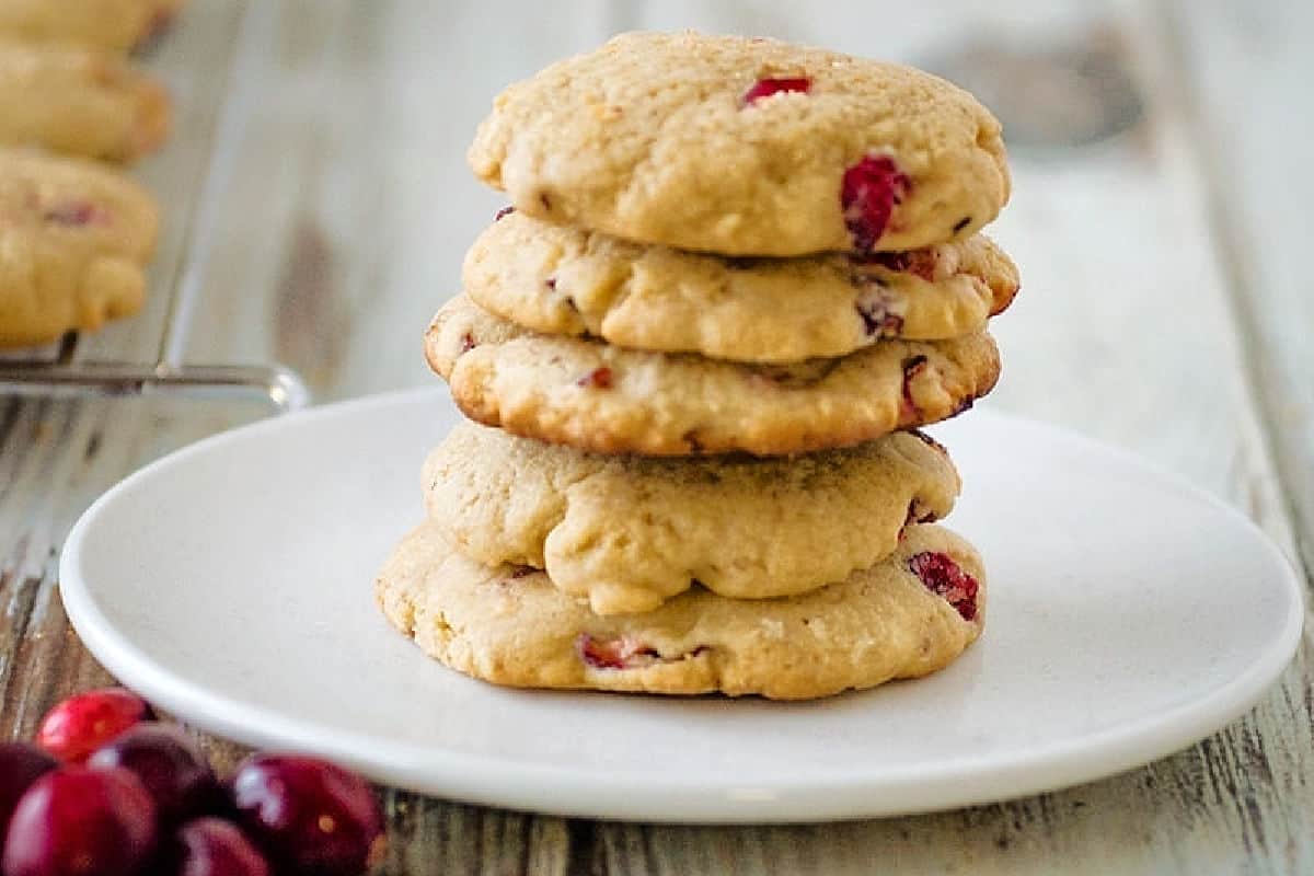 A stack of four cranberry cookies on a white plate, with a few cranberries beside the plate, on a wooden surface.