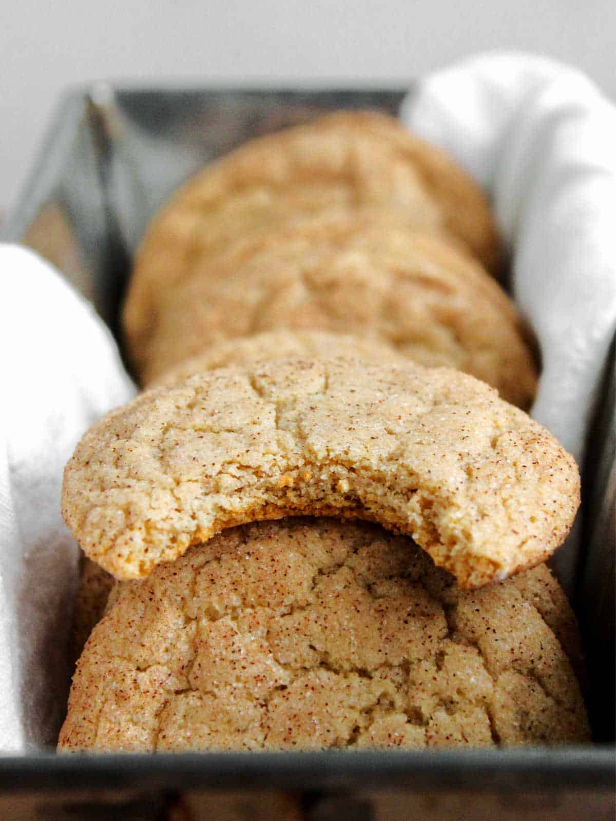 A row of freshly baked cookies, with the closest one having a bite taken out of it, resting in a tray lined with a white cloth.