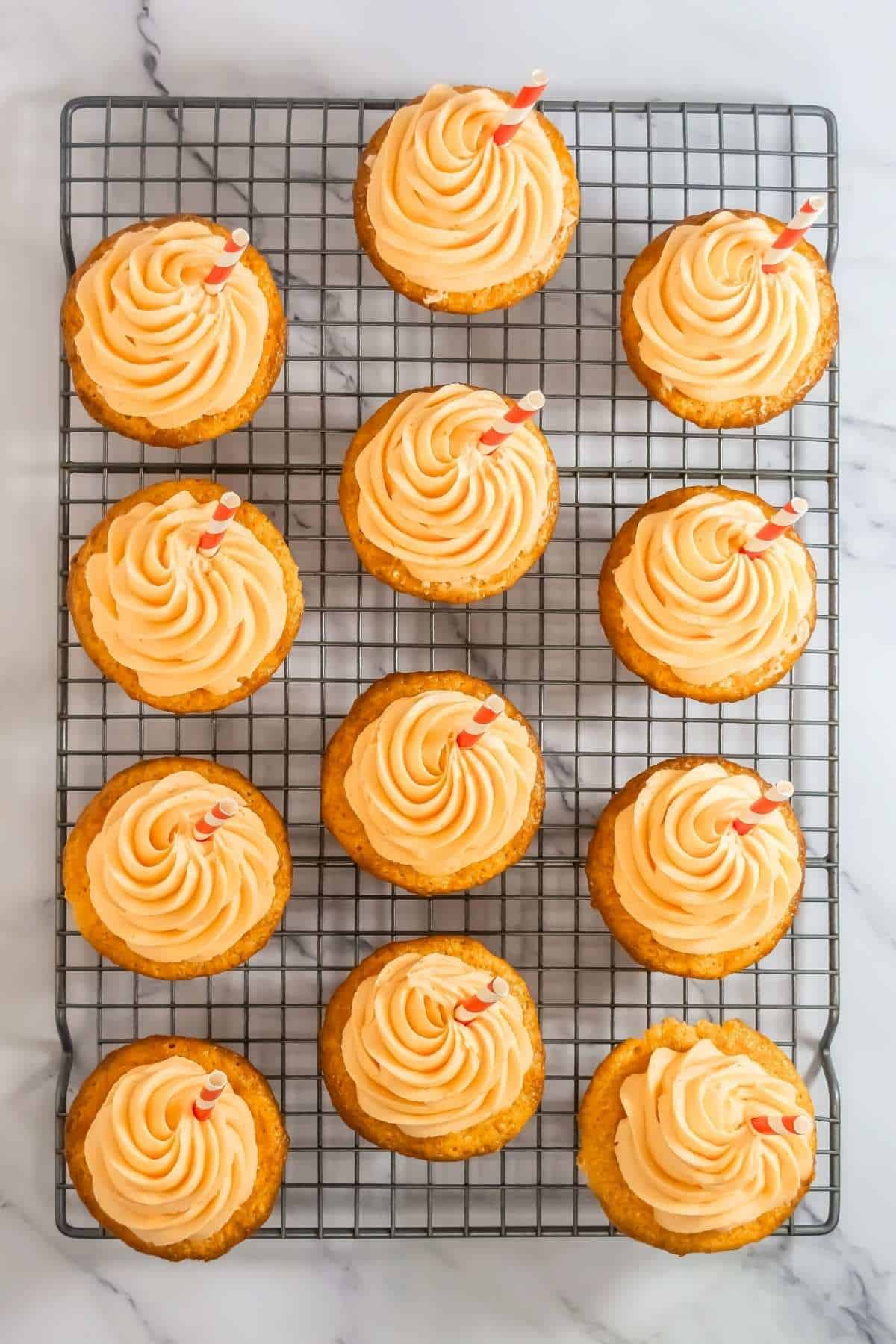 A dozen cupcakes with swirl frosting and striped straws arranged on a cooling rack atop a marble surface.