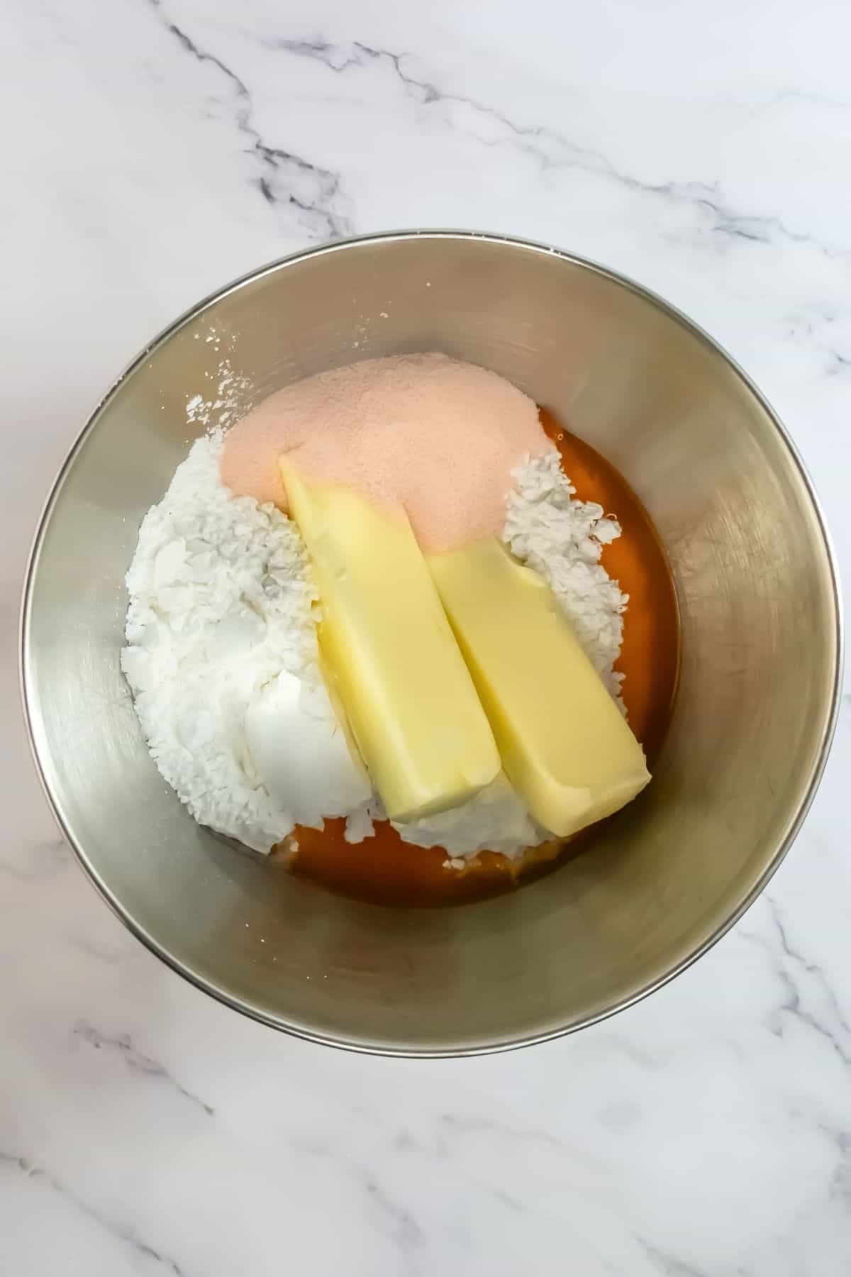 A metal mixing bowl containing flour, sugar, two sticks of butter, and a light brown substance, possibly vanilla extract, all on a marble countertop.