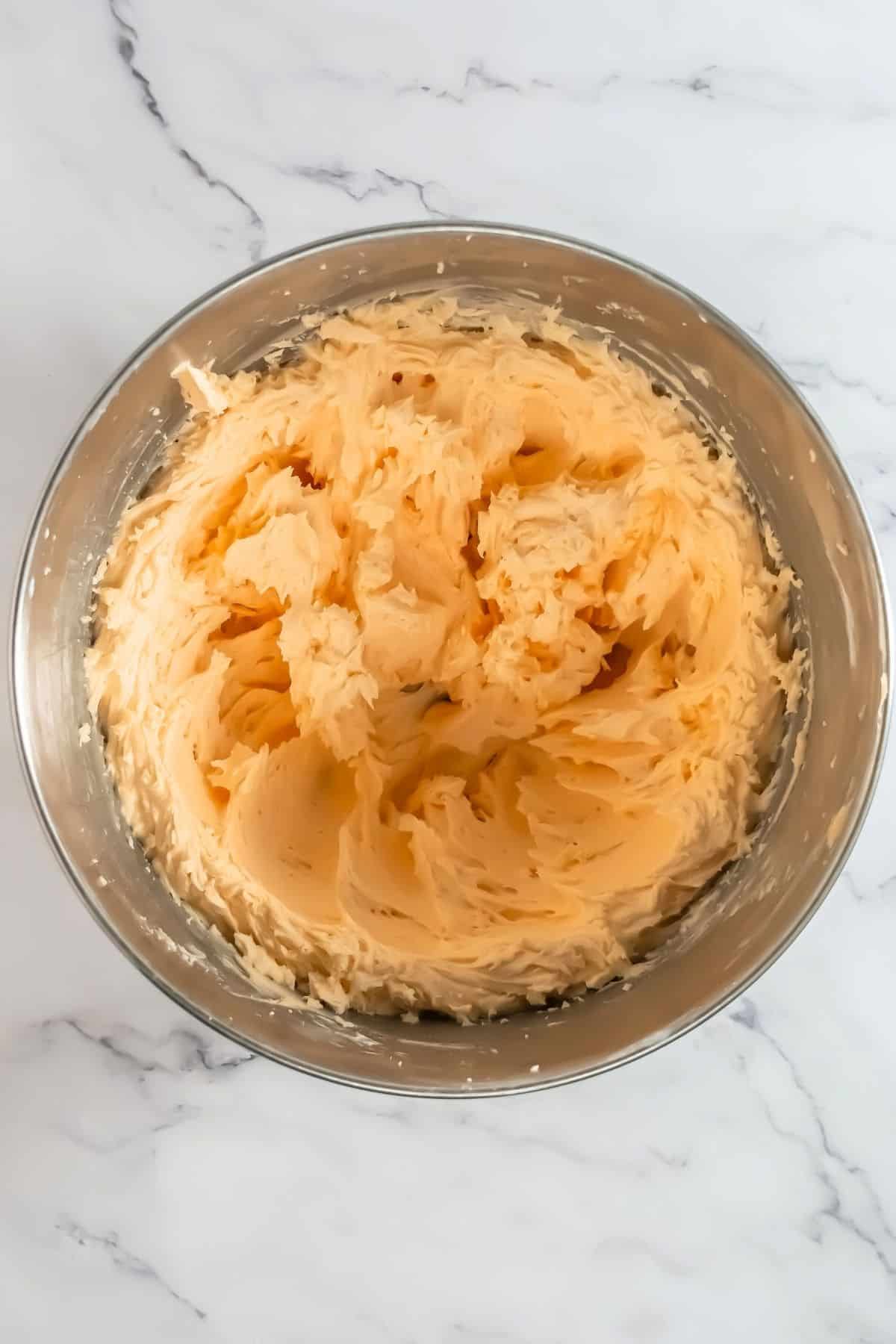 A metal mixing bowl containing creamy, yellow buttercream frosting on a white marble surface.