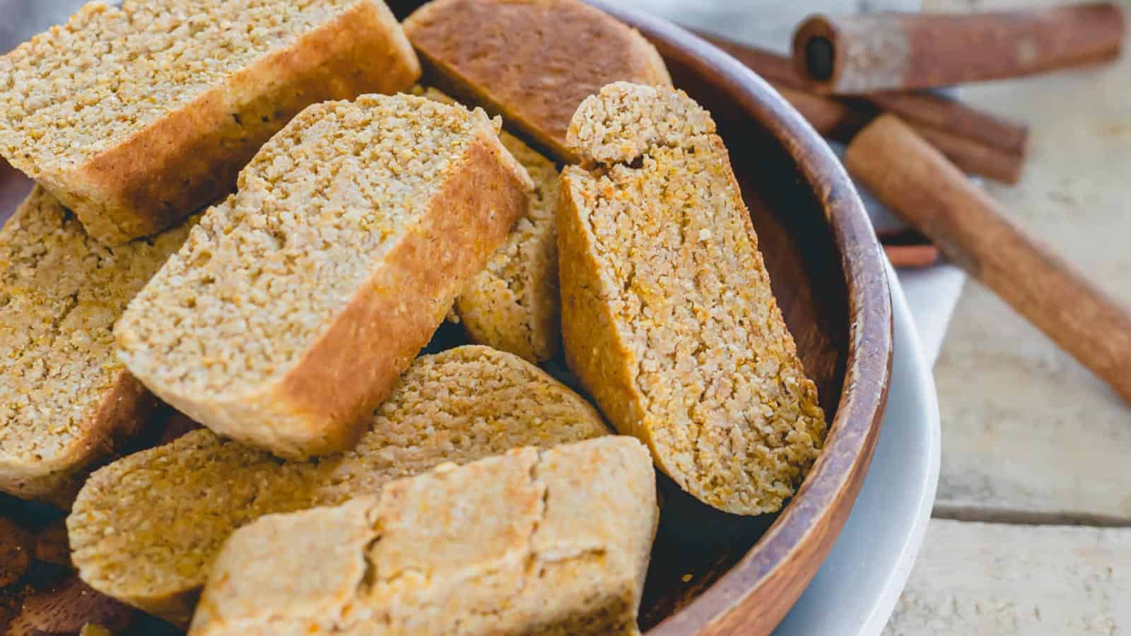 A wooden bowl filled with several coarse-textured, rectangular baked bread slices. Cinnamon sticks are placed in the background.