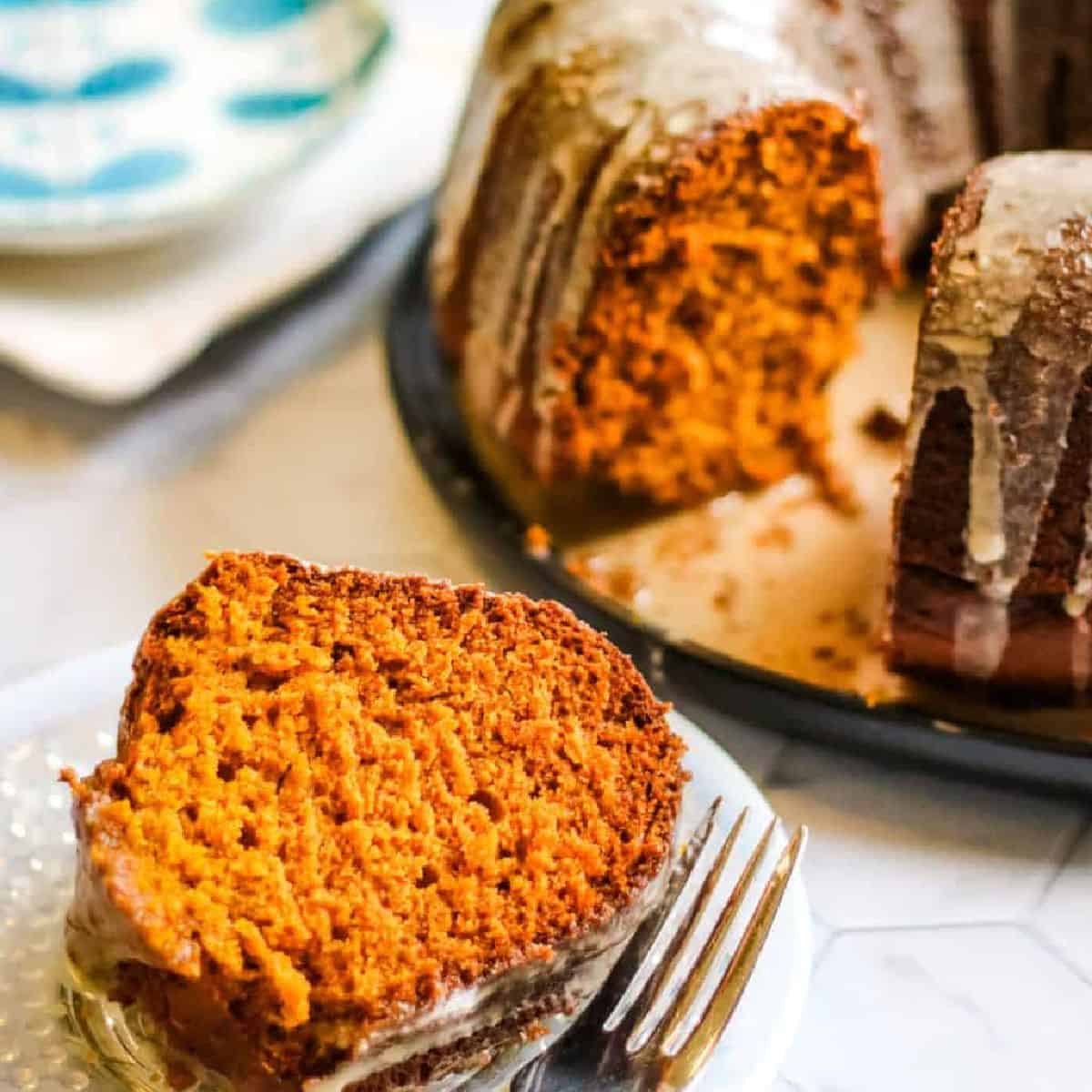 A slice of bundt cake with orange coloring and icing glaze sits on a white plate with a fork. The rest of the cake is visible in the background.