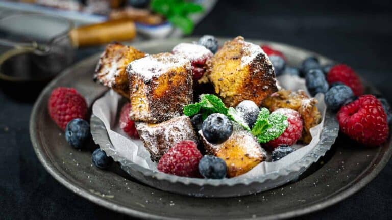 A plate with French toast cubes dusted with powdered sugar, garnished with blueberries, raspberries, and mint leaves.