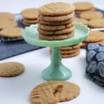 Stack of peanut butter cookies on a green cake stand, surrounded by more cookies on a table and cooling rack. A bitten cookie is in the foreground.