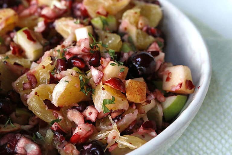 A close-up of a fruit salad with pineapple, apples, cherries, and pomegranate seeds in a white bowl.
