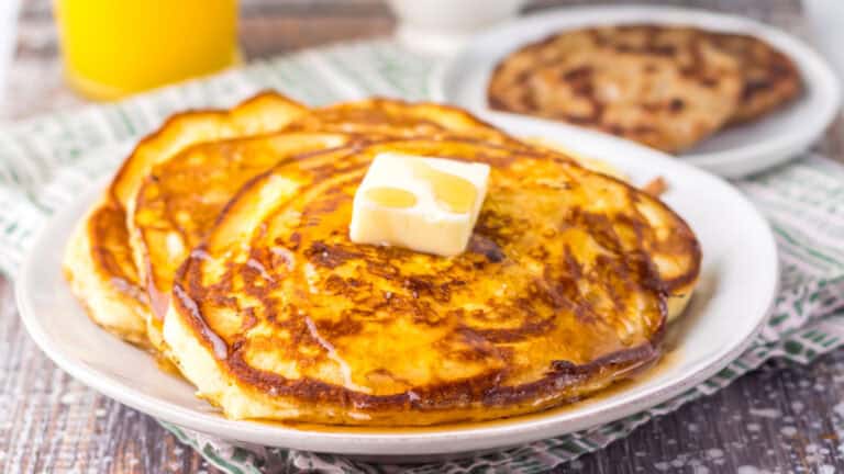 Stack of pancakes topped with a square of butter and syrup on a white plate, with a folded napkin underneath and a drink in the background.