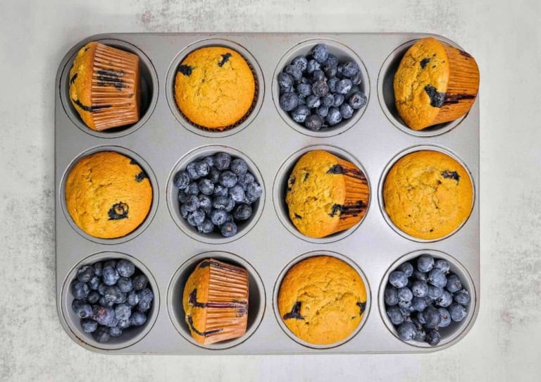 A muffin tin with six blueberry muffins and six compartments filled with fresh blueberries.