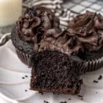 Three chocolate cupcakes with chocolate frosting are on a plate. One cupcake is cut in half to show its interior.