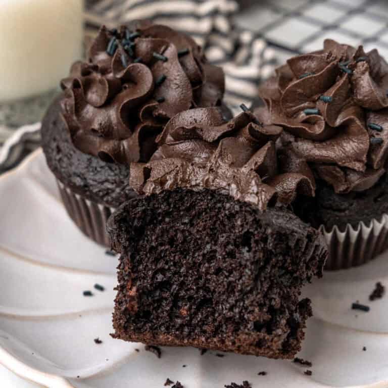 Three chocolate cupcakes with chocolate frosting are on a plate. One cupcake is cut in half to show its interior.