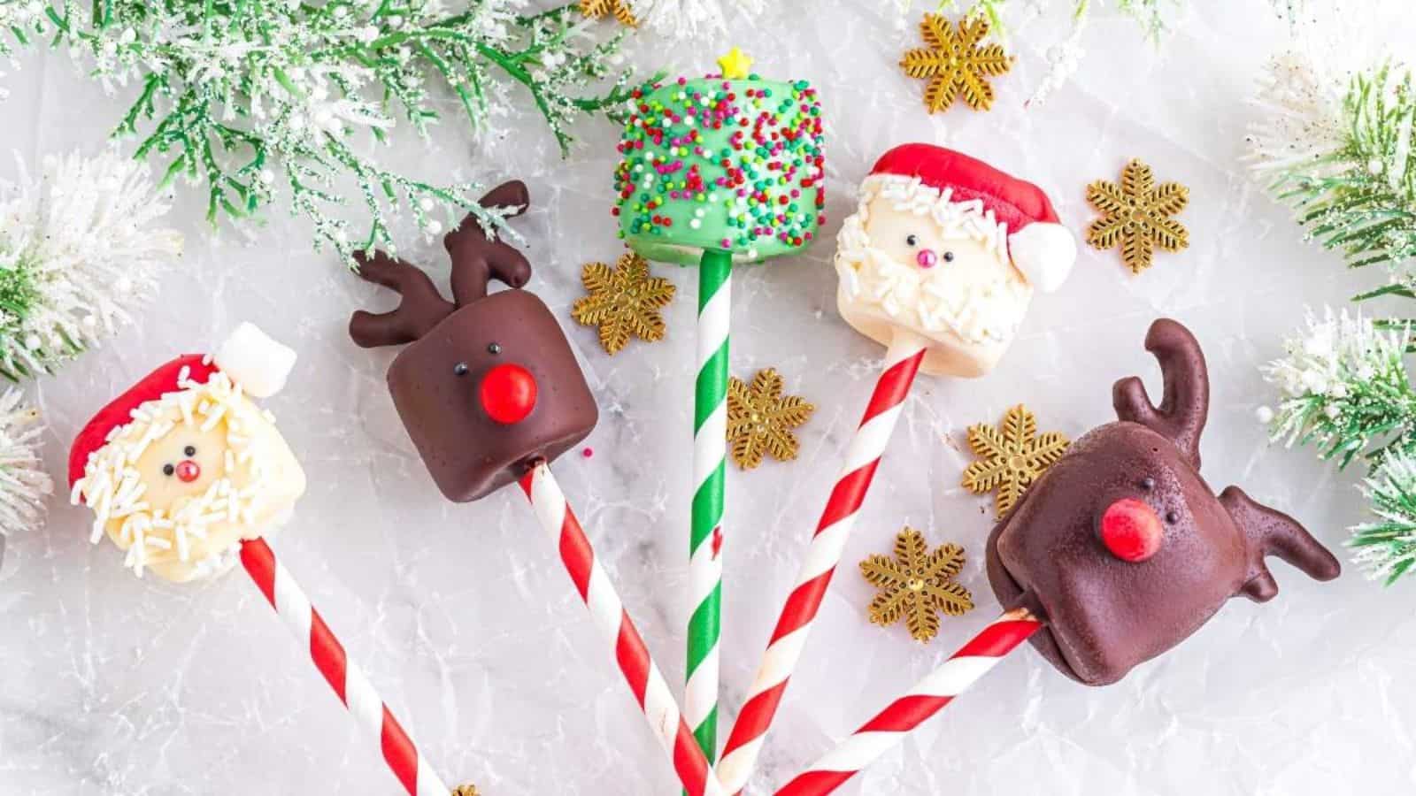 Five festive cake pops decorated as Santa and reindeer with red and white striped sticks, surrounded by snowflake decorations and greenery.