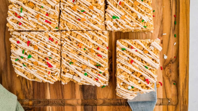A tray of nine rice crispy treats with white icing drizzle and red and green sprinkles, one being lifted with a spatula.