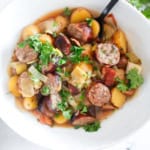 A white bowl of mixed stew with sliced sausages, potatoes, onions, and herbs, with a black spoon.