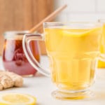 A glass mug of lemon ginger tea with a slice of lemon. A jar of honey and ginger root are in the background.