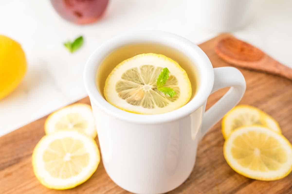 A white mug on a wooden surface filled with tea, topped with a lemon slice and mint. Slices of lemon and a wooden spoon are nearby.
