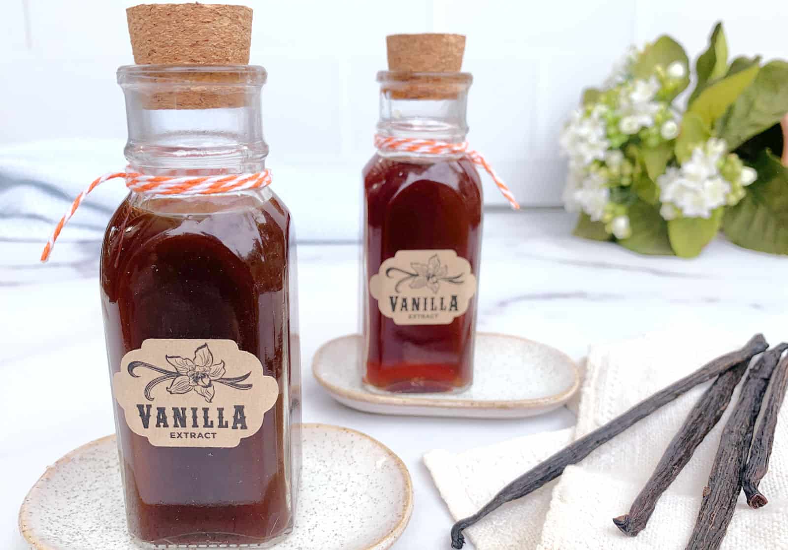 Two glass bottles of vanilla extract with cork stoppers and labels, placed on small plates. Vanilla pods and white towels are nearby, with green leaves and white flowers in the background.
