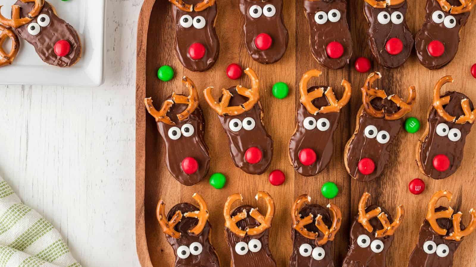 A tray of reindeer-themed chocolate cookies with pretzel antlers and red noses, decorated with candy eyes and M&Ms.