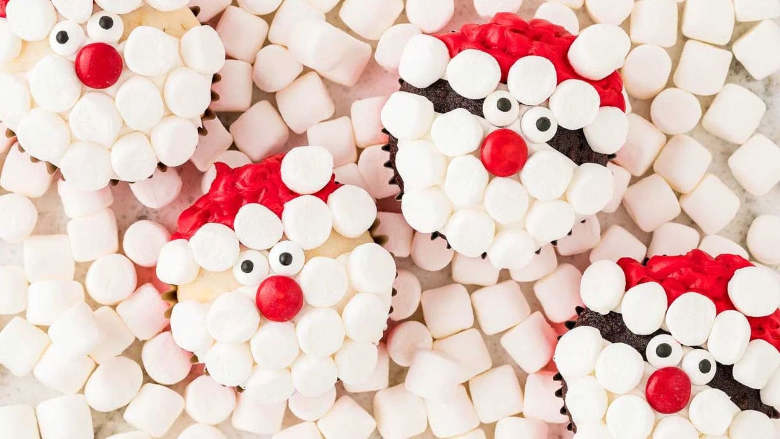 Cupcakes decorated as Santa faces with marshmallows and red frosting, surrounded by mini marshmallows.