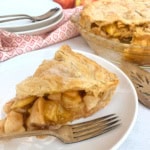 A slice of apple pie on a plate with a fork, next to a larger pie. Plates and a patterned cloth are in the background.