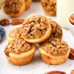 Close-up of six mini pecan tassies stacked on a white surface, surrounded by whole pecans.