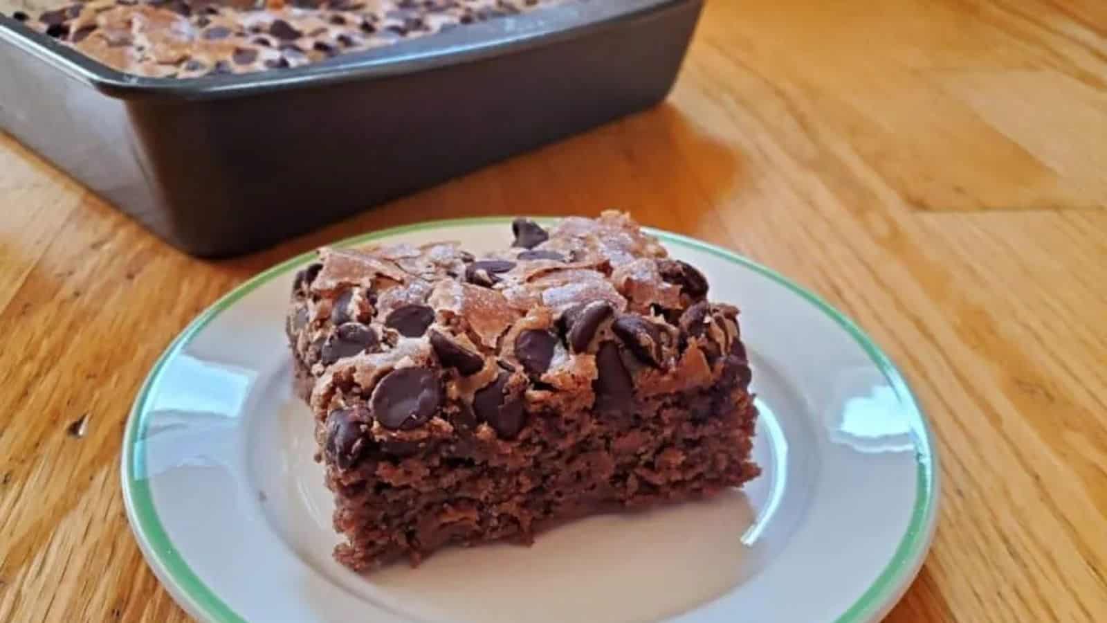 A small plate with a slice of chocolate chip applesauce cake with the pan holding the rest of the cake behind it.