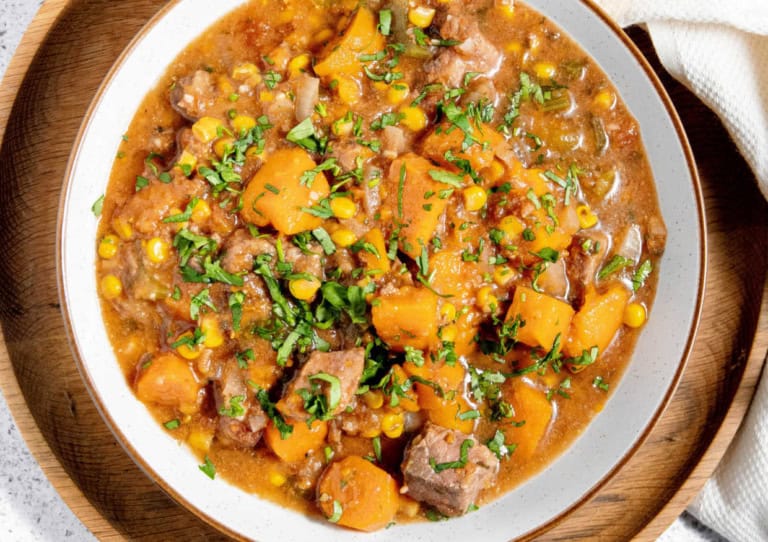 A bowl of hearty stew containing chunks of beef, corn, and orange vegetables garnished with fresh herbs.