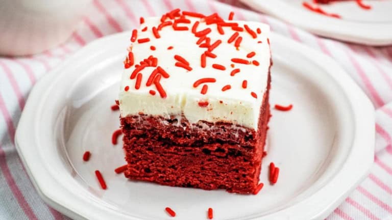 A slice of red velvet cake with cream cheese frosting and red sprinkles on a white plate.