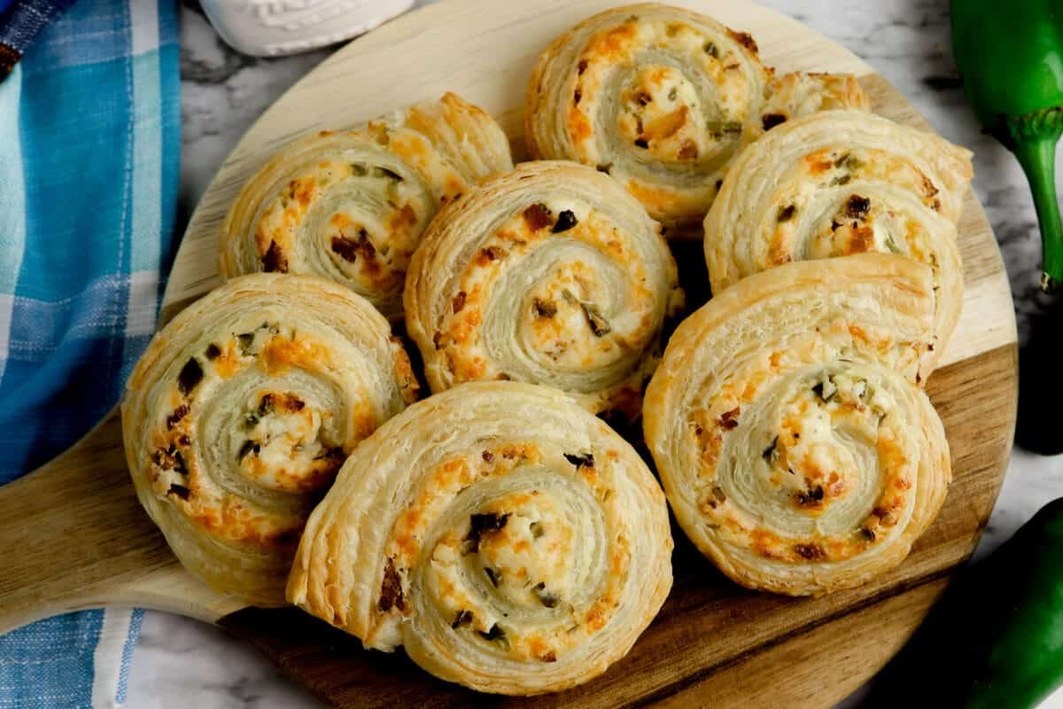 A wooden board with flaky, spiral-shaped pastries filled with cheese and herbs, surrounded by a checkered cloth and jalape&ntilde;os.