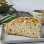 A slice of homemade bread on a white plate, with a green and white cloth and a beige mug in the background.