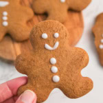 A hand holds a gingerbread cookie with a smiley face and three icing buttons. More cookies are on a wooden board in the background.