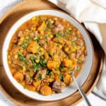 A bowl of stew with beef, potatoes, corn, and herbs, served on a wooden tray with a spoon and a white cloth.