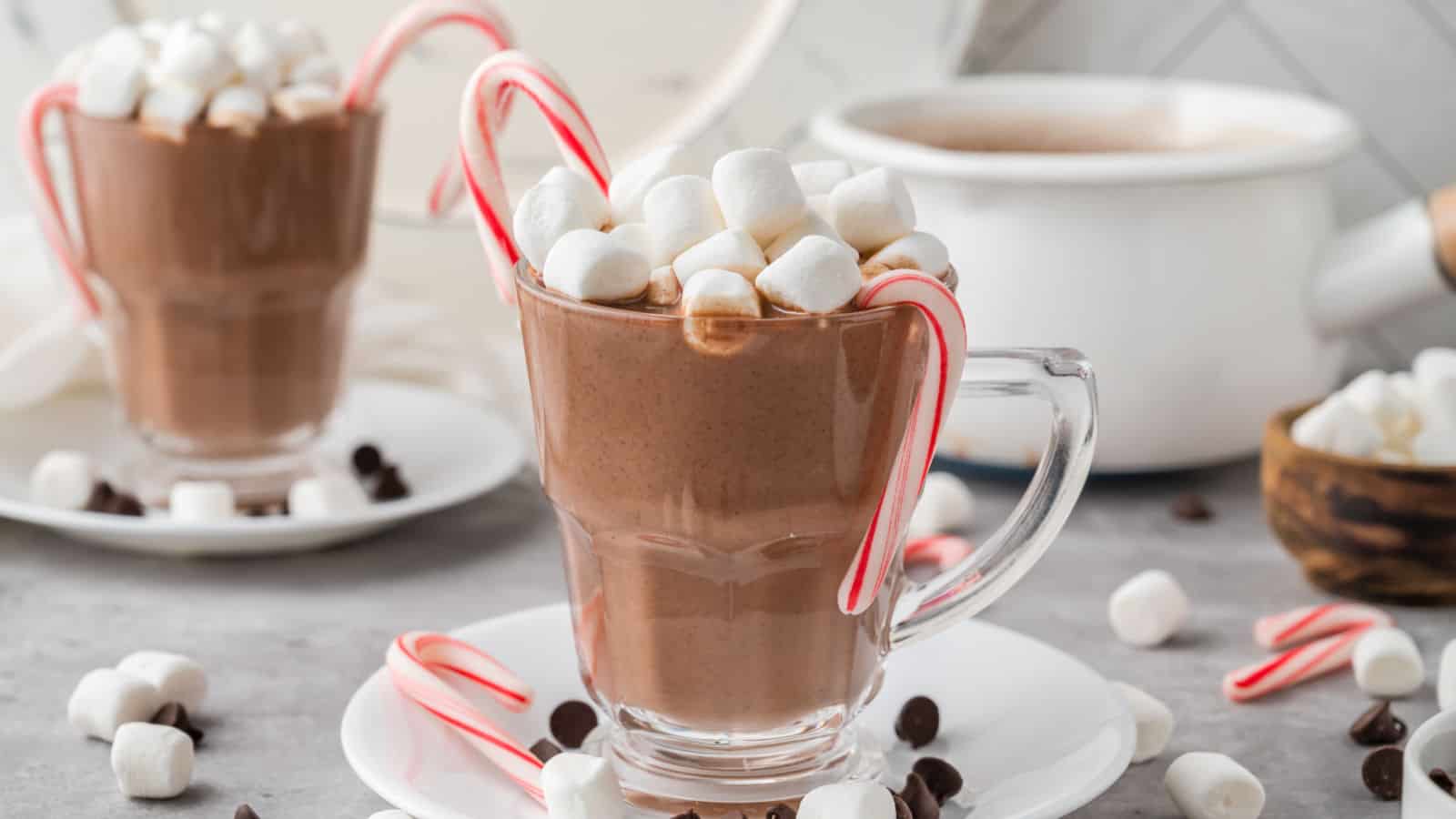 Two clear glass mugs of hot chocolate topped with marshmallows and candy canes on white plates, surrounded by more marshmallows and chocolate chips. A pot of hot chocolate is in the background.