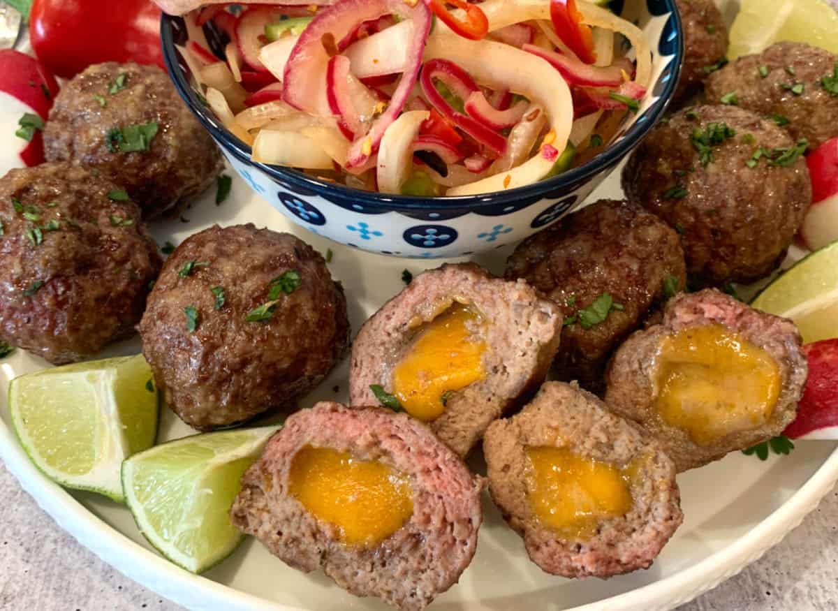 A plate of meatballs stuffed with melted cheese, garnished with lime wedges and herbs, surrounding a bowl of sliced onions and peppers.