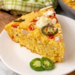 A slice of cornbread topped with jalapeños, tomatoes, and cheese on a white plate, with additional jalapeño slices on the side.