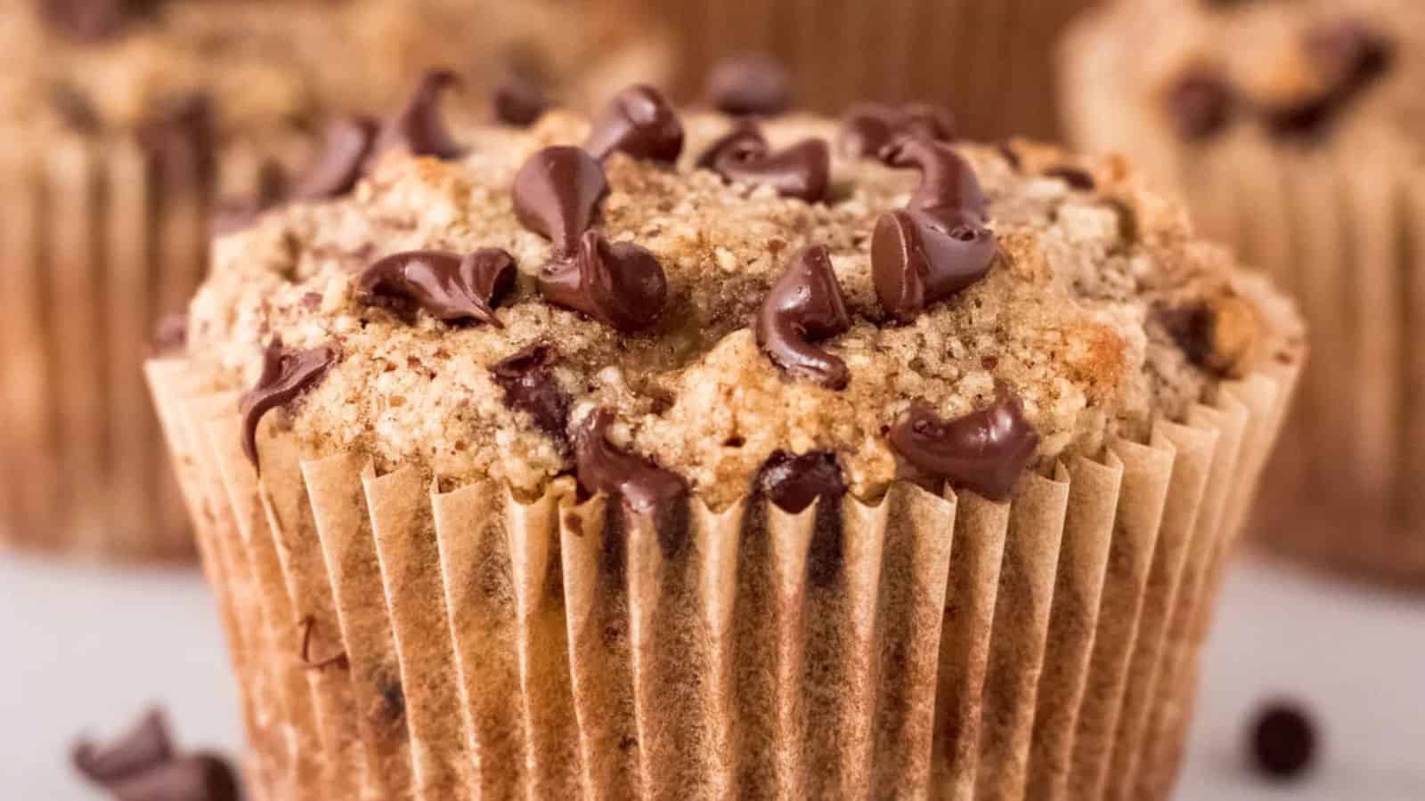 A close-up of a chocolate chip muffin in a paper liner, with crumbly topping and chocolate bits on the surface.