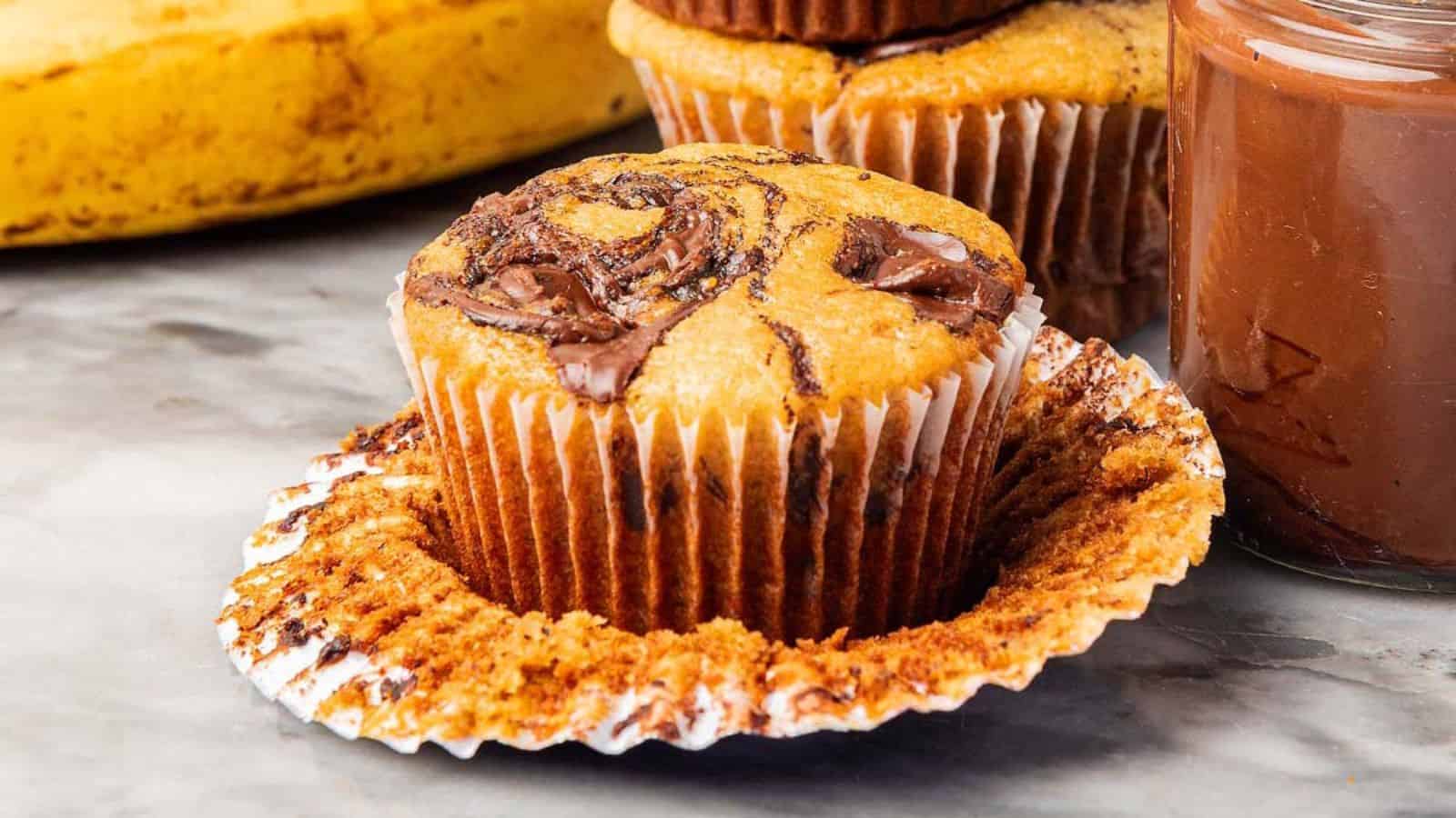 Close-up of a banana chocolate swirl muffin with the wrapper peeled back, surrounded by a jar of chocolate spread and a ripe banana.