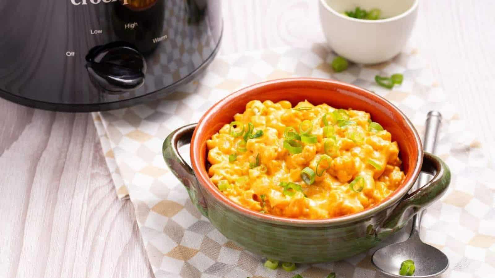 A bowl of cheesy macaroni and cheese topped with chopped green onions sits next to a slow cooker on a patterned cloth. A spoon is placed beside the bowl.