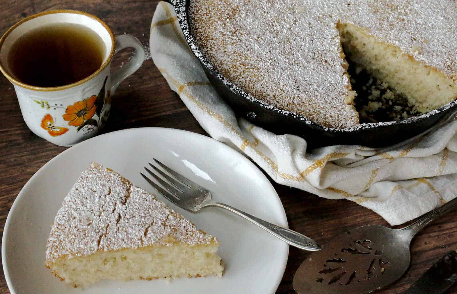 A slice of cake with powdered sugar on a plate with a fork, next to a cup of tea. A pan with the rest of the cake is in the background, sitting on a cloth.