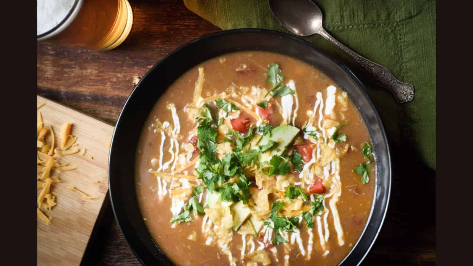 Bowl of soup topped with chopped tomatoes, avocado, cilantro, shredded cheese, and cream swirls. A spoon and green napkin are beside the bowl, with a glass of beer in the background.